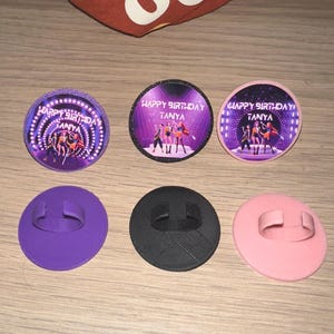 May include: A collection of six round party favors. Three are decorated with a stage scene and the text "HAPPY BIRTHDAY TANYA". The other three are solid colors: purple, black, and pink. Each has a loop on the back.