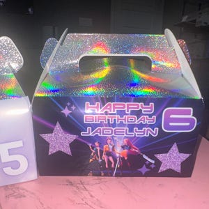 May include: A birthday gift box with a holographic top and handle. The front features the text "HAPPY BIRTHDAY JADELYN 6" with star and character graphics. The box is designed for a birthday celebration.