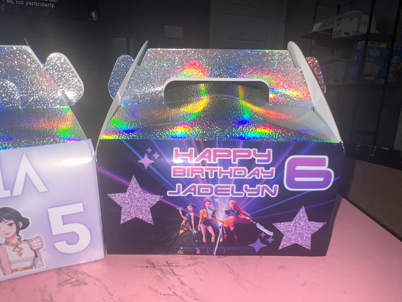 May include: A birthday gift box with a holographic top and handle. The front features the text "HAPPY BIRTHDAY JADELYN 6" with star and character graphics. The box is designed for a birthday celebration.