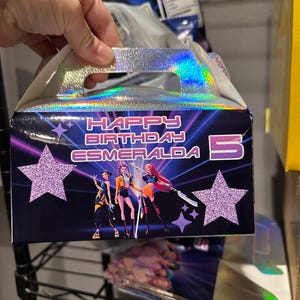 May include: A holographic birthday gift box with the text "HAPPY BIRTHDAY ESMERALDA 5". The box features a design with three figures, purple stars, and a holographic handle.