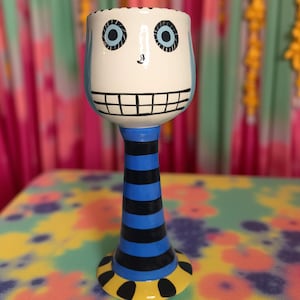 May include: A whimsical, hand-painted ceramic goblet with a face design. The goblet features a white bowl with blue accents, black eyes, and a smiling mouth. The stem is striped blue and black, with a yellow base adorned with black dots.