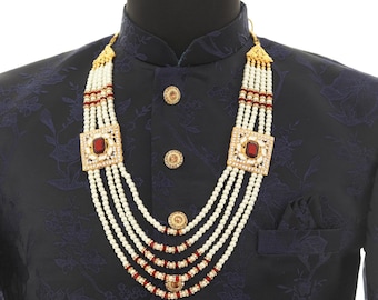 May include: A multi-strand pearl necklace with red and gold accents. The necklace features a square pendant with a red gemstone in the center. The necklace is worn on a black patterned shirt.