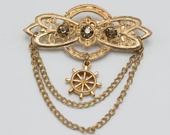 May include: A gold-toned brooch with a delicate filigree design and three sparkling rhinestones. The brooch features a dangling chain with a small gold wheel charm.