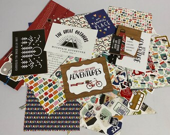 May include: A collection of travel-themed scrapbook paper, stickers, and embellishments. Includes designs with mountains, bicycles, and the phrases "The Great Outdoors," "Fill Your Life With Adventures," and "Let the Adventure Begin." Various colors and patterns.