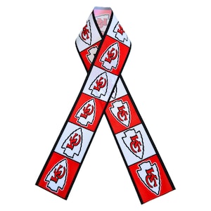 May include: A ribbon-shaped scarf featuring a repeating pattern of red and white squares. Each square displays a red arrowhead logo with the letters "KC" in white. The scarf has a black border and is displayed against a white background.