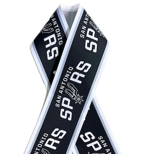 May include: Black and white ribbon featuring the text "SAN ANTONIO SPURS" and the team logo. The ribbon has a white border and is folded into a loop. The Spurs logo is a stylized spur with a star.