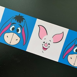 May include: Ribbon featuring alternating blue and white panels with cartoon character faces. The ribbon showcases Eeyore and Piglet from Winnie the Pooh. The ribbon is ideal for crafting, gift wrapping, or adding a touch of whimsy to various projects.