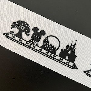 May include: White ribbon with a repeating black silhouette design featuring a castle, a tree with an elephant, a Mickey Mouse head, and a geometric structure. The ribbon has a ribbed texture and is angled across a black surface.