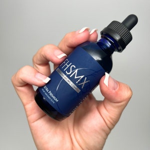 May include: A dark blue glass bottle with a black dropper is held in a hand. The bottle has a navy blue label with the text "FHSHMX Cellular Balancing Complex" and "Ultra Premium Dietary Supplement 60ml". The bottle is filled with a dark blue liquid.