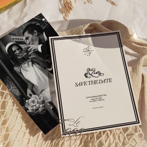 May include: A black and white photograph and a cream-colored "Save the Date" card. The card has a black border and features swan illustrations and the text "SAVE THE DATE". The photograph shows a couple toasting with champagne. The card includes the wedding date and location.