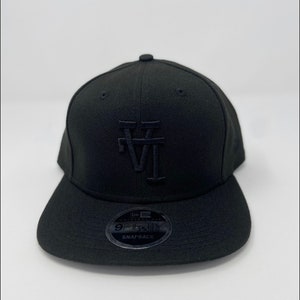 May include: A black fitted baseball cap with a black embroidered "LA" logo on the front. The cap has a black adjustable snap closure and a black New Era logo on the back.