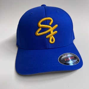 May include: A royal blue baseball cap with a gold embroidered letter 'S' on the front. The cap has a black and silver sticker on the side that says "The Original Flex Fit" and "L/XL It Fits Your Life."