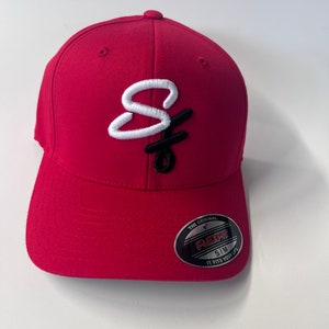 May include: A red baseball cap with a curved brim. The cap features a white and black embroidered logo on the front. The Flexfit label is visible on the side. The cap is a size S/M.