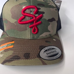 May include: A camouflage baseball cap with a red embroidered "SF" logo. The cap has a black mesh back and a black "The Authentic Yupoong Snapback Yupoong Since 1974" label.