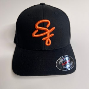 May include: Black baseball cap with an orange embroidered "SF" logo. The cap has a black Flexfit label on the back.