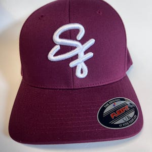 May include: A maroon baseball cap with a white embroidered "S" logo. The cap has a Flexfit label on the side, indicating it is a size L/XL.