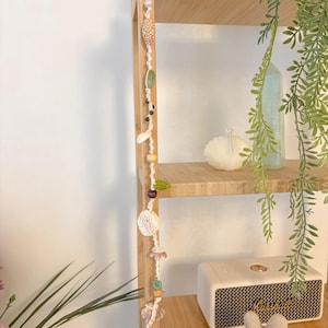 May include: A cream macrame wall hanging with shells, beads, and natural elements. The design includes wooden beads, green leaves, and pink and clear crystal-like beads. The hanging is displayed on a wooden shelf. The shelf also holds a speaker and decorative items.