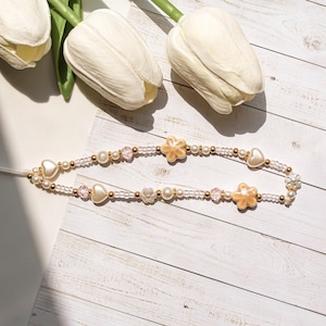 Peut inclure: Une lanière de téléphone ornée de perles blanches, roses et dorées. Elle comprend des perles en forme de cœur et de fleur, ainsi que de petites perles et des perles transparentes. Posée sur une surface en bois blanc, avec des fleurs blanches.