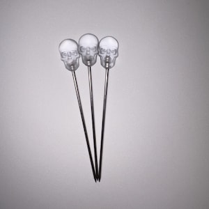 May include: Three clear skull-shaped cocktail picks with silver stems. The skull heads are detailed with eye sockets, a nose cavity, and teeth. The picks are arranged in a triangular formation against a gray background, suitable for Halloween or gothic-themed events.
