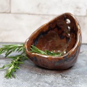 May include: A small, handmade ceramic bowl with a rustic, organic shape. The bowl is glazed in shades of brown and has dark blue accents. Fresh rosemary sprigs are placed inside the bowl. The bowl is displayed on a gray surface.
