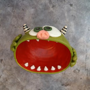 May include: A whimsical, green monster-shaped bowl with a wide-open red mouth. The bowl features white teeth, green eyes, a pink nose, and striped horns. The bowl is designed to hold small items.