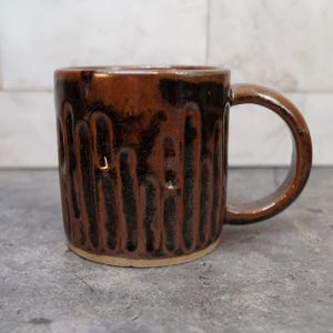 May include: A brown ceramic mug with a dark brown and black textured design. The mug has a rounded handle and a light-colored base. The exterior features vertical, textured lines, creating a unique, handcrafted appearance. This mug is suitable for coffee, tea, or other beverages.