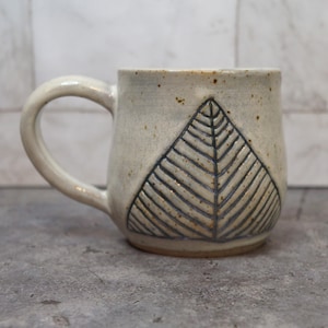 May include: A handmade ceramic mug with a cream-colored glaze and a large, dark blue leaf design. The mug has a rounded shape, a sturdy handle, and a speckled texture. The mug is suitable for hot or cold beverages.