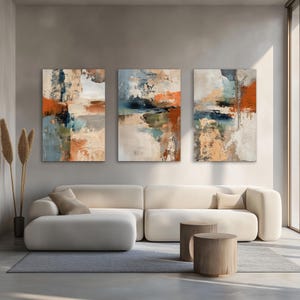 May include: Three abstract art pieces with a modern design. The paintings feature a mix of colors, including orange, blue, and beige, with textured brushstrokes. The artwork is displayed above a cream-colored sofa in a well-lit room.