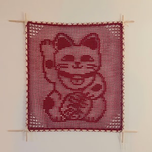 Puede incluir: Un tapiz de pared rojo tejido a ganchillo con un dise&ntilde;o de gato de la suerte. El gato est&aacute; saludando con su pata, un s&iacute;mbolo de buena suerte. La obra de arte cuadrada est&aacute; enmarcada con clavijas de madera e hilo blanco.