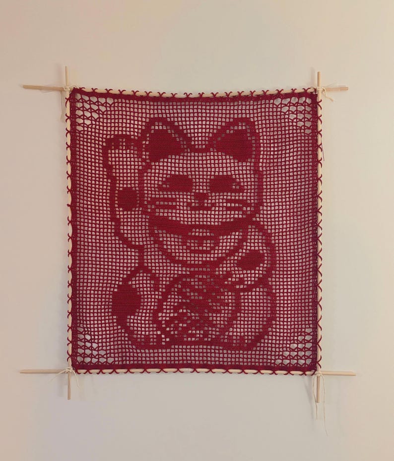 Puede incluir: Un tapiz de pared rojo tejido a ganchillo con un dise&ntilde;o de gato de la suerte. El gato est&aacute; saludando con su pata, un s&iacute;mbolo de buena suerte. La obra de arte cuadrada est&aacute; enmarcada con clavijas de madera e hilo blanco.