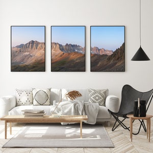 May include: Three framed landscape photographs of a mountain range with a blue sky and clouds. The photos are hung on a white wall above a white couch with pillows and a throw blanket. A black butterfly chair is in the foreground.