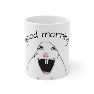 Op de afbeelding: Een witte keramische mok met een zwarte lijntekening van een lachend knaagdier met een roze neus en twee opvallende voortanden. Boven de illustratie staat de tekst "good morning" gedrukt in een eenvoudig, zwart, kleingeschreven lettertype.