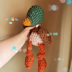 May include: A handmade crochet duck toy with a green head, orange beak, and brown and white body. The duck has orange feet and is made with a chunky yarn. This soft toy is perfect for children.
