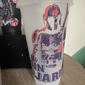 May include: A clear plastic reusable cup with a design of a Mandalorian character from the Star Wars franchise. The design is in shades of gray, black, and pink. The text "Din Djarin" is printed on the cup.