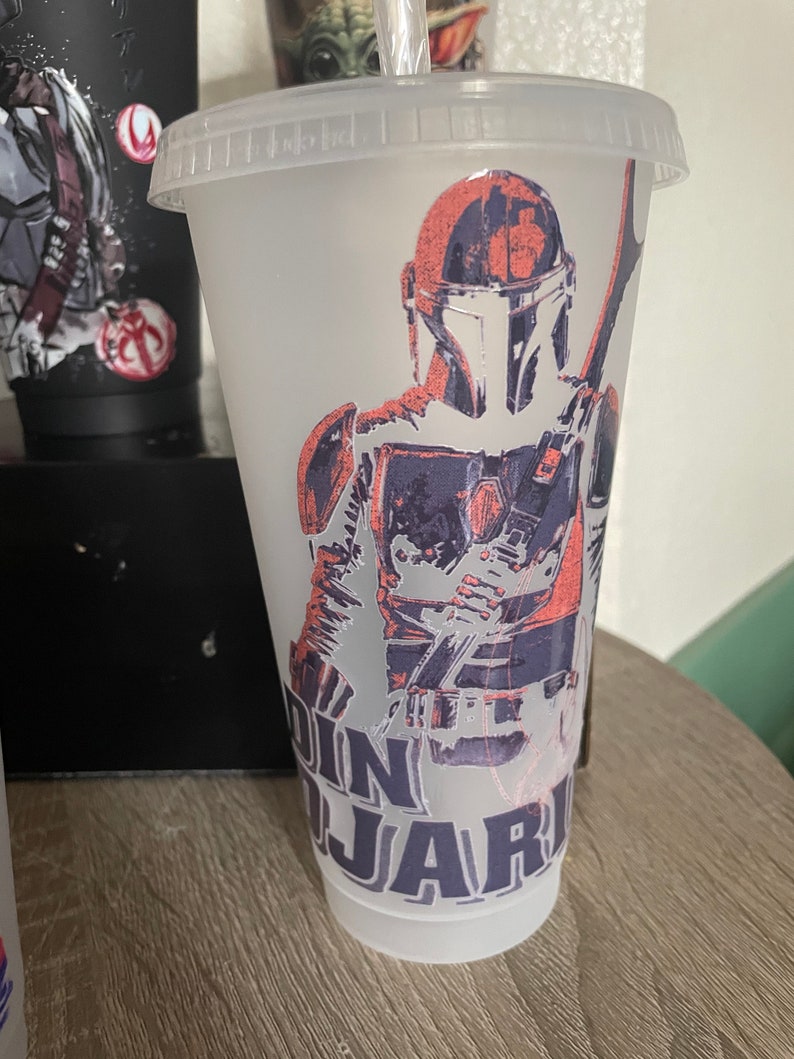 May include: A clear plastic reusable cup with a design of a Mandalorian character from the Star Wars franchise. The design is in shades of gray, black, and pink. The text "Din Djarin" is printed on the cup.