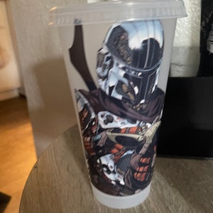 May include: A clear plastic tumbler with a graphic of a Mandalorian character from Star Wars. The character is wearing a helmet and holding a weapon. The tumbler is sitting on a wooden surface.
