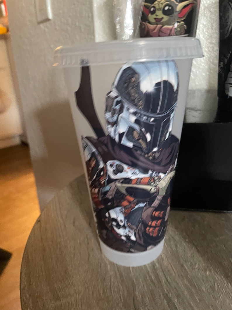 May include: A clear plastic tumbler with a graphic of a Mandalorian character from Star Wars. The character is wearing a helmet and holding a weapon. The tumbler is sitting on a wooden surface.