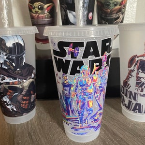 May include: Five clear plastic cups with different Star Wars themed designs. The cups are all the same size and shape. The designs include a Mandalorian, Baby Yoda, and a group of Star Wars characters. The text "STAR WARS" is printed on one of the cups.
