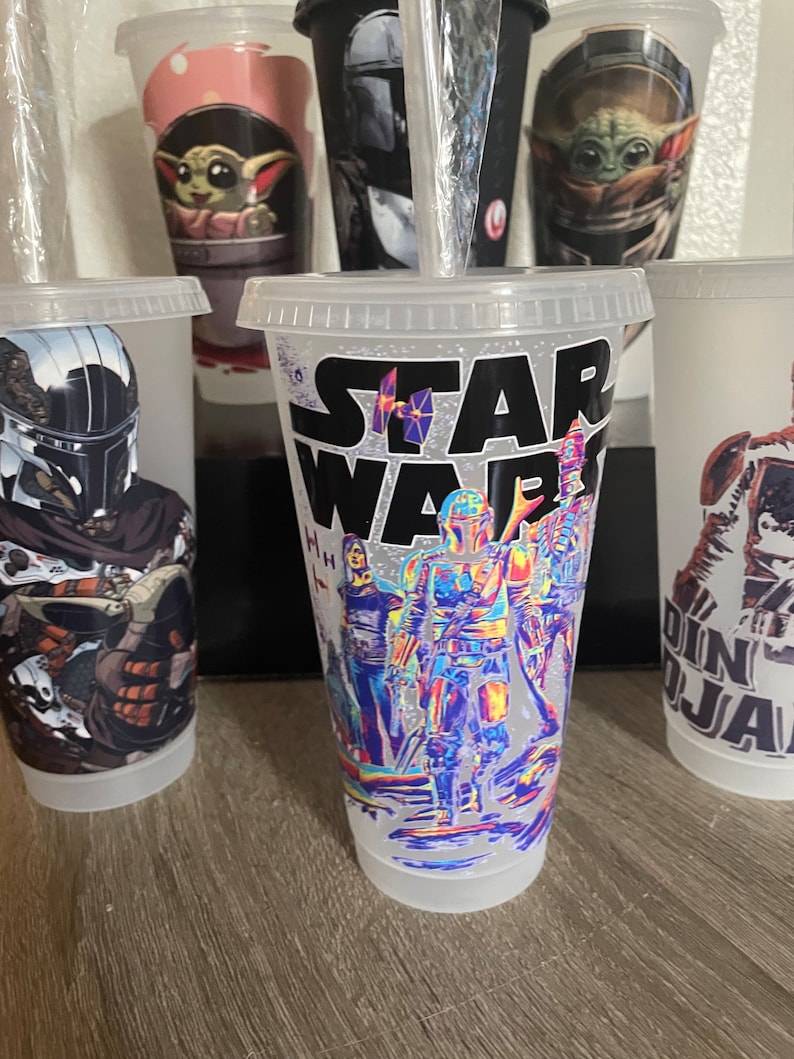 May include: Five clear plastic cups with different Star Wars themed designs. The cups are all the same size and shape. The designs include a Mandalorian, Baby Yoda, and a group of Star Wars characters. The text "STAR WARS" is printed on one of the cups.