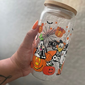 May include: A clear glass tumbler with a bamboo lid, featuring a Halloween-themed design. The design includes cartoon characters dressed in skeleton costumes, pumpkins, bats, and a spiderweb. The tumbler is designed for cold beverages.