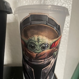 May include: A clear plastic cup with a green and brown illustration of a character from the Star Wars franchise. The character is wearing a helmet and has large eyes. The cup has a straw.