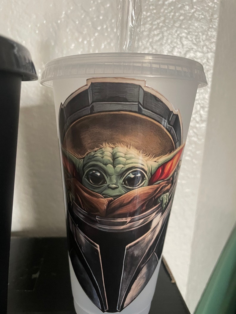 May include: A clear plastic cup with a green and brown illustration of a character from the Star Wars franchise. The character is wearing a helmet and has large eyes. The cup has a straw.