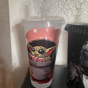 May include: A white reusable cup with a pink, gray, and green design featuring a cartoon character from the Star Wars franchise. The character is sitting in a pod and has large ears and green skin.