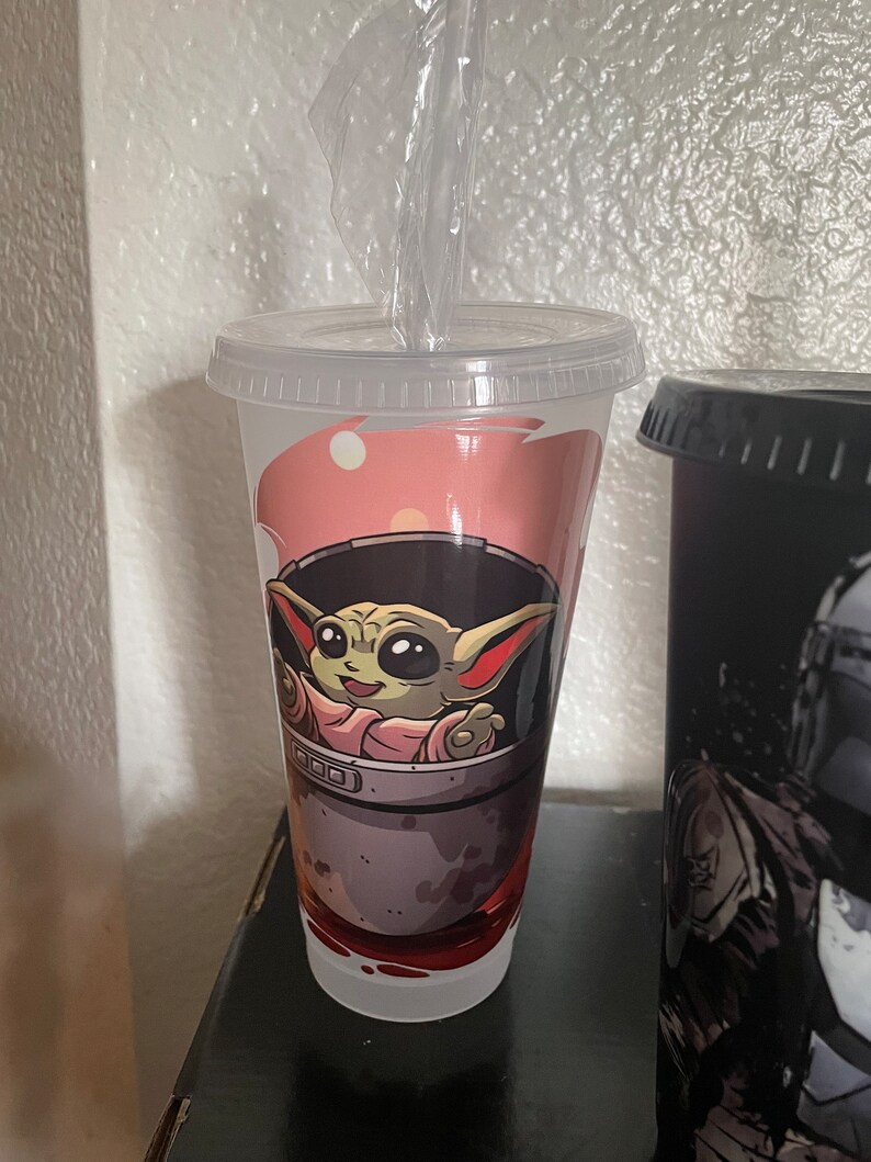 May include: A white reusable cup with a pink, gray, and green design featuring a cartoon character from the Star Wars franchise. The character is sitting in a pod and has large ears and green skin.
