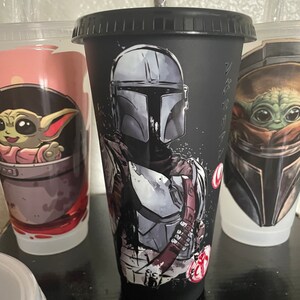 May include: Black reusable cup with a graphic of a Mandalorian character from Star Wars. The cup has a black lid and a straw.