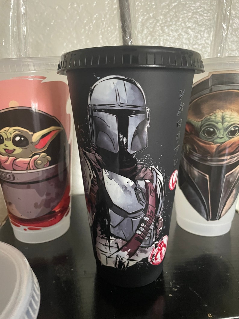 May include: Black reusable cup with a graphic of a Mandalorian character from Star Wars. The cup has a black lid and a straw.