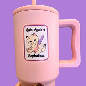 Puede incluir: Un termo rosa con asa y tapa. El termo presenta una pegatina con el texto "Cats Against Capitalism" y una ilustraci&oacute;n de un gato sosteniendo un cuchillo. El termo es sostenido por una mano con esmalte de u&ntilde;as oscuro.