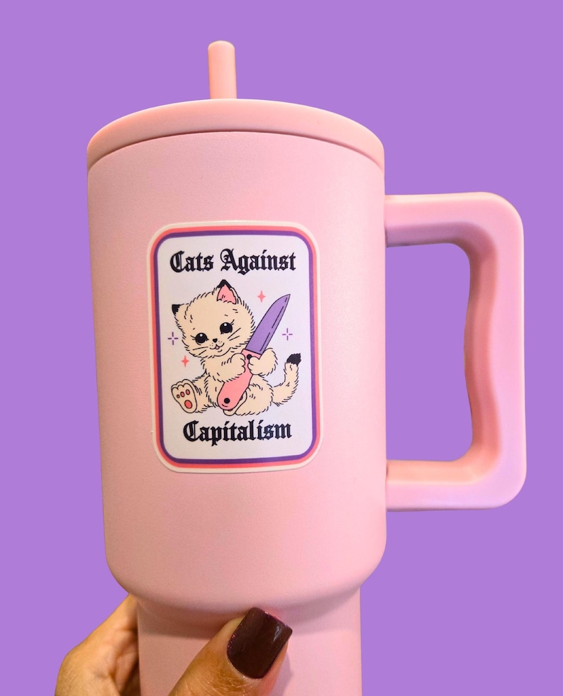 Puede incluir: Un termo rosa con asa y tapa. El termo presenta una pegatina con el texto "Cats Against Capitalism" y una ilustraci&oacute;n de un gato sosteniendo un cuchillo. El termo es sostenido por una mano con esmalte de u&ntilde;as oscuro.