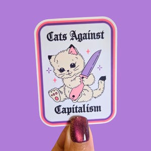 Puede incluir: Una pegatina con un gato de dibujos animados sosteniendo un cuchillo rosa. El texto "Cats Against Capitalism" se muestra arriba y abajo de la ilustraci&oacute;n del gato. La pegatina tiene un fondo blanco con un borde rosa y morado.
