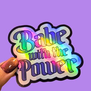 May include: A holographic sticker with the text "Babe with the Power" in a retro font. The sticker has a black outline and a rainbow gradient effect. The background is a solid purple color.